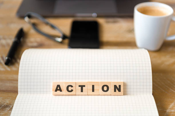 istock Closeup on notebook over wood table background, focus on wooden blocks with letters making Action text 989180878