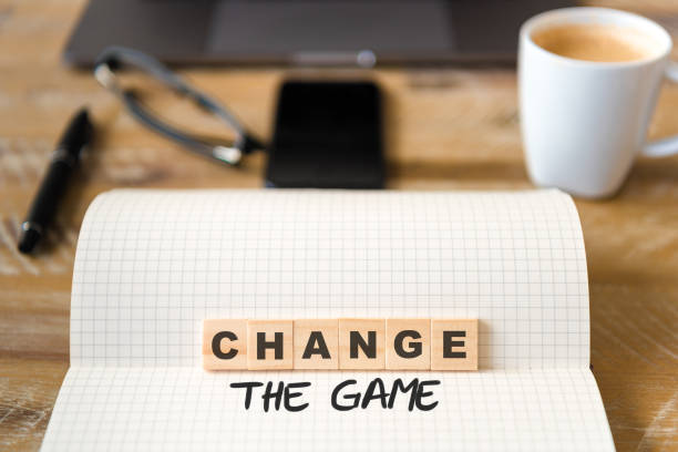 istock Closeup on notebook over wood table background, focus on wooden blocks with letters making Change the Game text 989181854