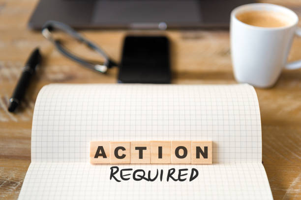 istock Closeup on notebook over wood table background, focus on wooden blocks with letters making Action Required text 989184320