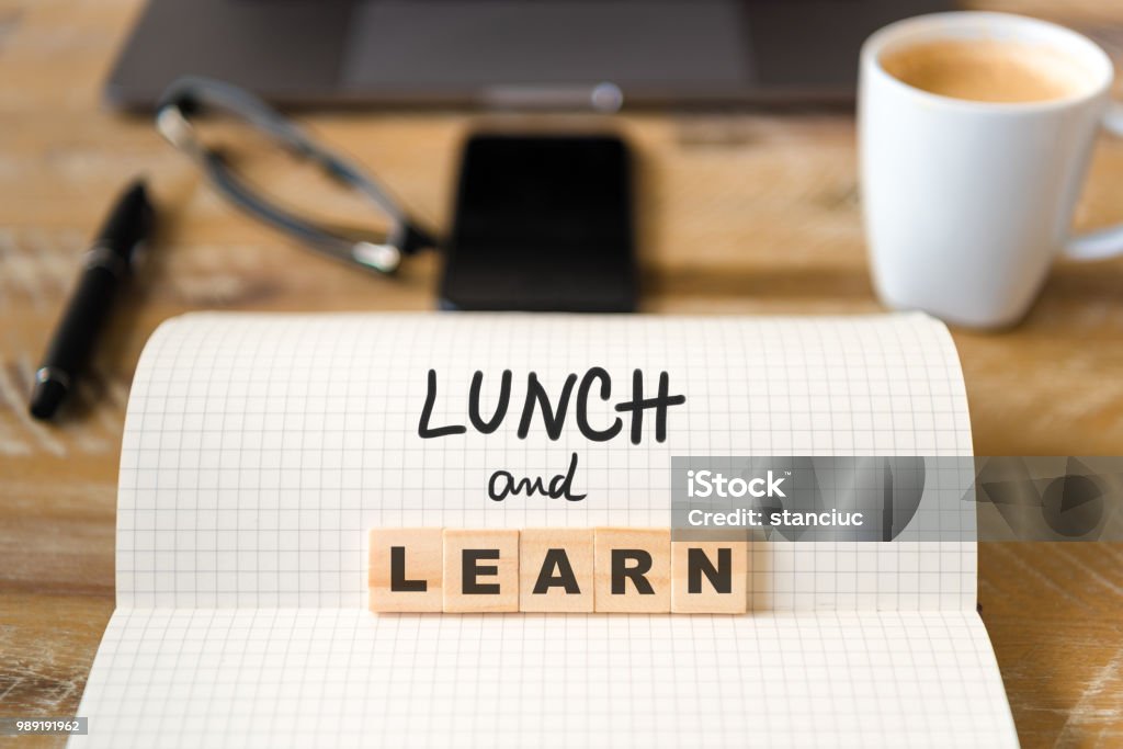 Closeup on notebook over wood table background, focus on wooden blocks with letters making Lunch and Learn text Closeup on notebook over wood table background, focus on wooden blocks with letters making Lunch and Learn text. Concept image. Laptop, glasses, pen and mobile phone in defocused background Lunch Stock Photo