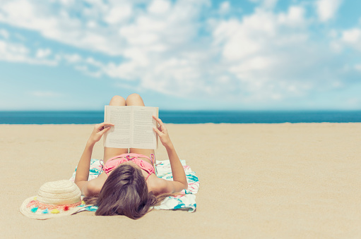 Young brunette woman lying on the beach relaxing reading a book with the sea and horizon in the background on summer day travel and tourism concept