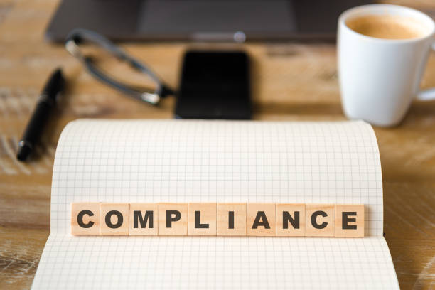 istock Closeup on notebook over wood table background, focus on wooden blocks with letters making Compliance text 989194564