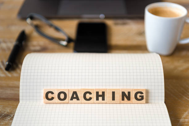 istock Closeup on notebook over wood table background, focus on wooden blocks with letters making Coaching text 989198692