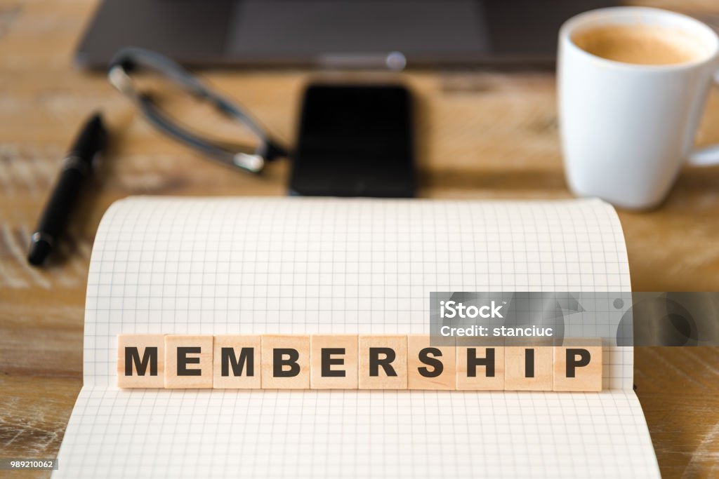 Closeup on notebook over wood table background, focus on wooden blocks with letters making Membership text Closeup on notebook over wood table background, focus on wooden blocks with letters making Membership text. Concept image. Laptop, glasses, pen and mobile phone in defocused background Organized Group Stock Photo