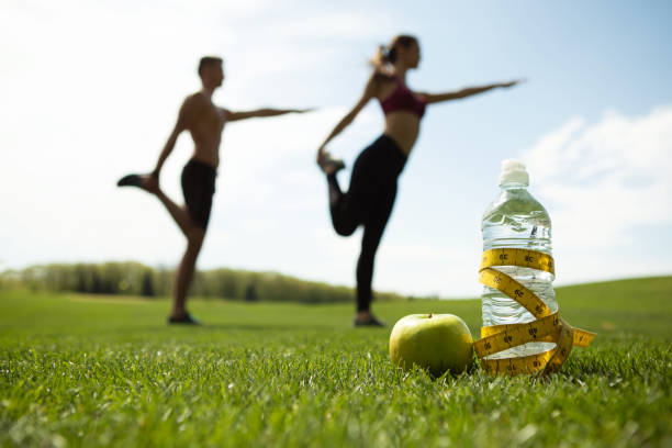 Man and woman are training in green park Focus on bottle of water with centimeter band and apple on grass. Sporty couple is doing stretching exercises together on lawn. They are standing on one foot and bending other while keeping balance centimeter photos stock pictures, royalty-free photos & images