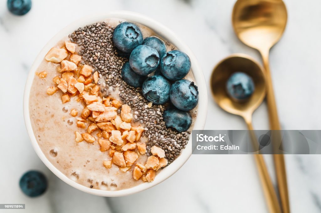 Top view of sa moothie bowl with fresh ripe blueberry, nuts, chia, banana and soya milk over white background. The concept of healthy eating and vegetarian food. Smoothie Stock Photo