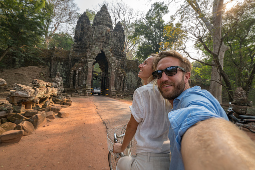 Shot of a carefree young couple taking a selfie while riding a bicycle, ancient temples in Angkor Wat temple complex. People travel concept.\nCambodia