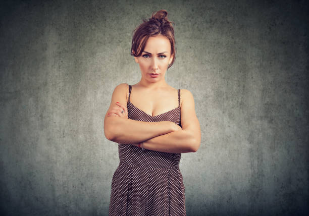 Young woman in dress holding hands crossed and looking at camera with anger and complaint on gray background