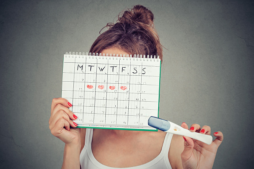mujer ocultando detrás de un calendario de períodos y mostrando una prueba de embarazo positiva photo