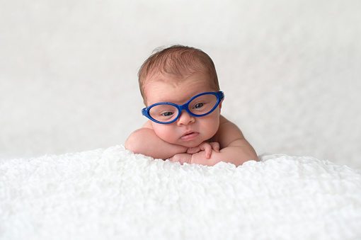 Portrait of nine day old, newborn baby girl. She is wearing blue, cat eye glasses and is looking at the camera.