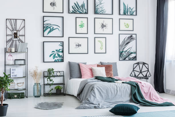 istock Gallery with simple posters hanging on the wall in bright bedroom interior with many pillows on bed, fresh plants and plastic chair standing next to window with drapes 989795610