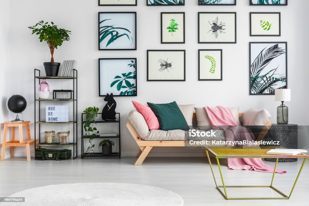 Gold Metal Table Standing In White Living Room Interior With Decor On Black  Metal Racks Fresh Green Plants Light Grey Sofa With Cushions And Pink  Blanket And Posters Hanging On The Wall