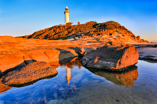 Shallow tide puddle with sandstone rocks on headland plato of Norah head with Norah lighthouse in warm morning light against blue clean sky.
