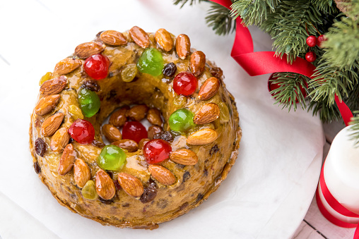 Glazed round hollow colorful Christmas fruitcake topped with almonds and glace cherries on white plate with green pine needles and red ribbon decorations