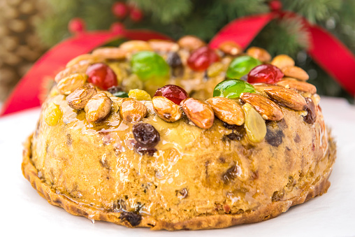 Close up of glazed round hollow colorful Christmas fruitcake topped with almonds and glace cherries on white plate with green pine needles and red ribbon decorations in background