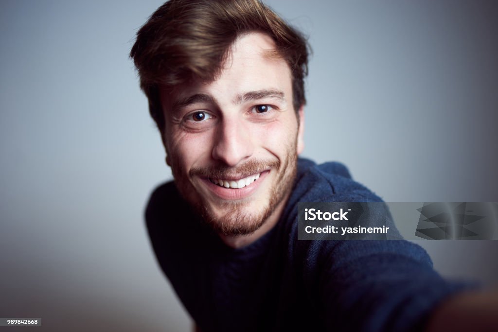 Mignon jeune homme regardant la caméra et le sourire dans l’atelier. Vraies personnes prenant un autoportrait seul avec un visage heureux - Photo de Hommes libre de droits