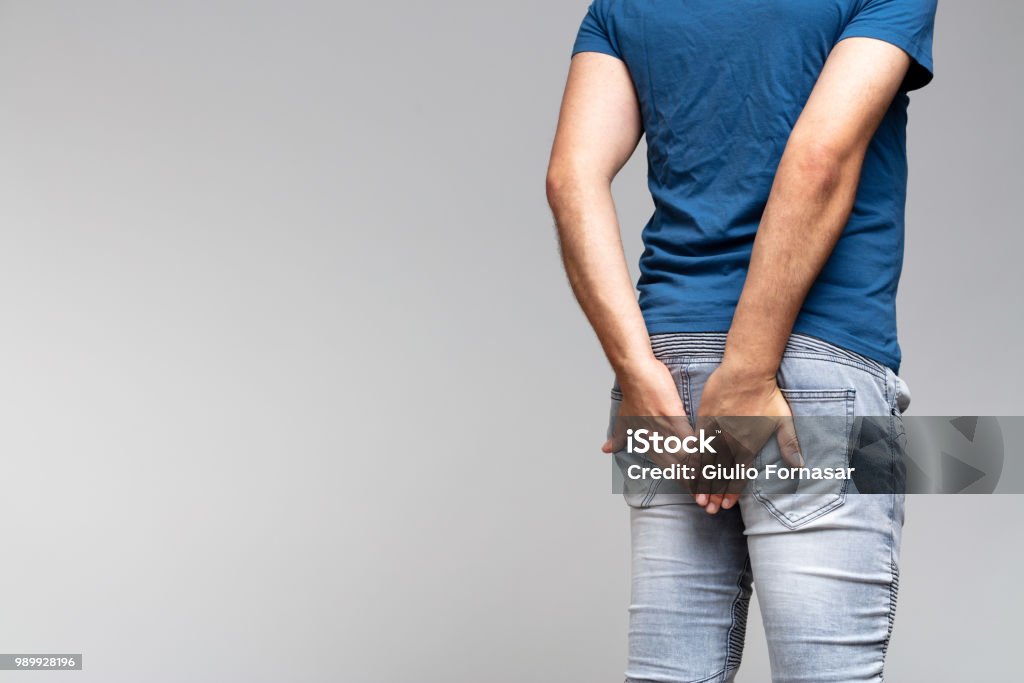 caucasian man holding his buttocks in denim jeans caucasian man in light blue t-shirt holding his buttocks in denim jeans on a gray background with copy space. Buttocks Stock Photo