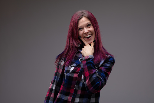 Fun playful attractive woman laughing at the camera with her hand to her chin and modern purple tinted long hair over a grey studio background