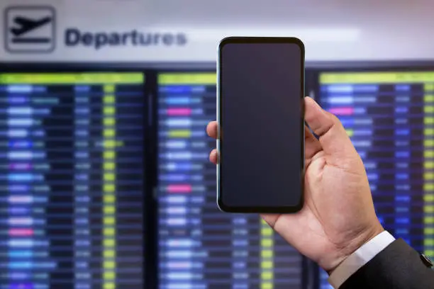 Smartphone Mockup Image. Businessman holding Mobile Phone on Hand in front of Departures Board to check Flight Information. Blank Display Screen as Clipping Path