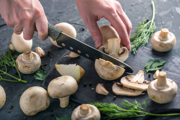 istock Women's hands are cutting champignons with a knife on the dark kitchen table. Cooking mushroom dishes. Close up 990104802