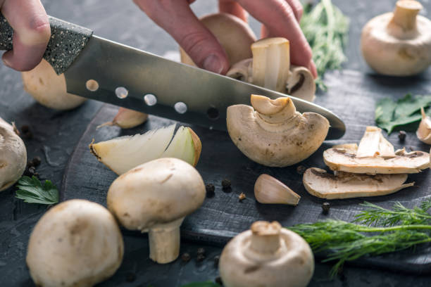 istock Women's hands are cutting champignons with a knife on the dark kitchen table. Cooking mushroom dishes. Close up 990104812