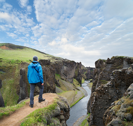 Tourist in blue jacket stands on top of a hill. Fjadrargljufur canyon in south east Iceland with the Fjadra river flowing through it. Amazing attraction. Summer day with clouds