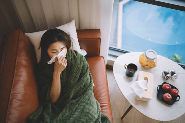 an sick asian chinese female lying on sofa in living room covered with blanket and wiping nose with tissue paper an sick asian chinese female lying on sofa in living room covered with blanket and wiping nose with tissue paper Fever Treatment stock pictures, royalty-free photos & images