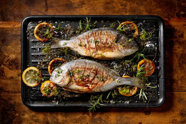 Grilled  fish, sea bream, dorada with the addition of spices, herbs and lemon on the grill plate located on a wooden background,  top view. Healthy eating concept