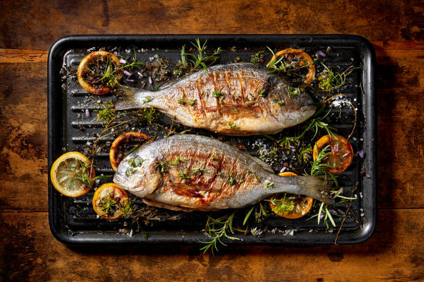 grilled  fish, sea bream, dorada with the addition of spices, herbs and lemon on the grill plate located on a wooden background,  top view. - prepared fish seafood barbecue grilled imagens e fotografias de stock