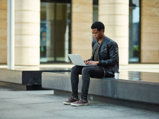 istock African American student in leather jacket sitting on stone bench outdoors and using laptop for studying 990304062