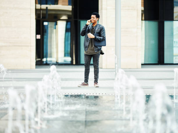 istock Smiling young man in black leather jacket holding disposable coffee cup and laptop and chatting on cell phone on city street near fountain 990304132