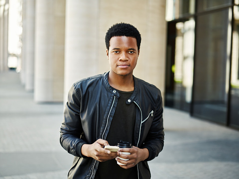 Portrait of serous African American guy looking at camera while walking down street with paper coffee cup and cell phone in his hands