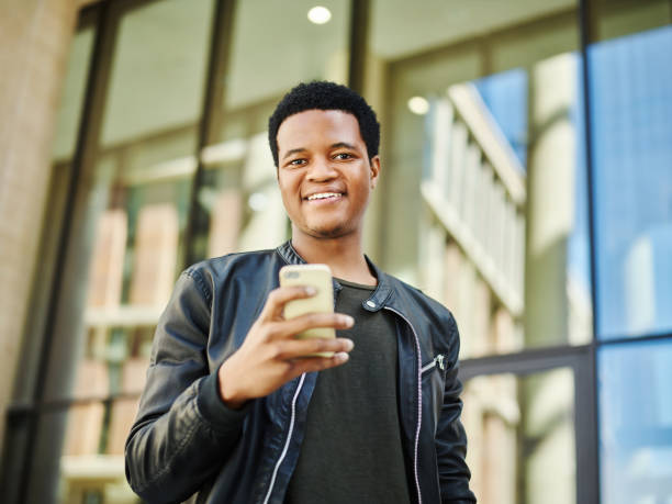 cintura-up retrato de feliz joven afro americano mirando a cámara con smartphone en la mano. chico con navegación internet en teléfono al aire libre - waistup fotografías e imágenes de stock