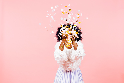 Celebrating happiness, young woman  throwing confetti obscuring her face, isolated on pink background