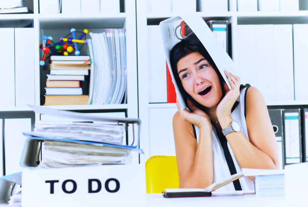 istock Funny overworked girl with a folder on her head sitting in the workplace cluttered with folders. Reaction of a subordinate when the boss orders her to do too much 990340310