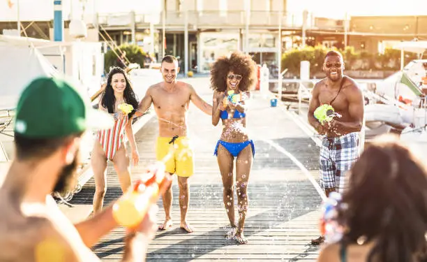 Multiracial happy friends having crazy fun with waterfight battle at summer location - Carefree vacation concept with young people millennials using water gun at beach docks - Warm backlight filter