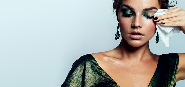 beautiful young girl with a bright make-up and in a shiny green dress striatet makeup from her face with a wet napkin. Hairstyle - curls are gathered in a bun.