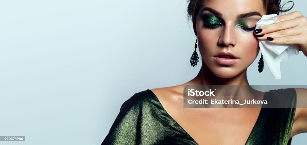 A girl is washing cosmetics. beautiful young girl with a bright make-up and in a shiny green dress striatet makeup from her face with a wet napkin. Hairstyle - curls are gathered in a bun. Make-Up Stock Photo