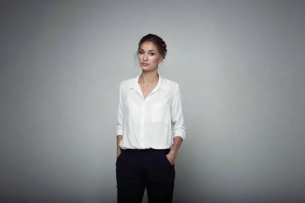 Interested businesswoman with trendy makeup posing on gray background in studio. Indoor photo of serious young lady in white blouses classic black pants standing in confident pose