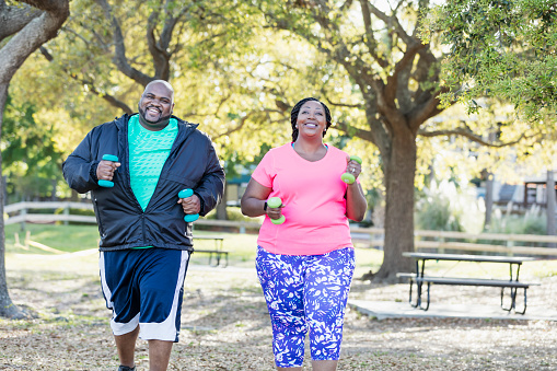 An African-American couple in the park, exercising together. They are lifting hand weights, power walking, smiling and moving toward the camera. They are both plus size models with large builds.