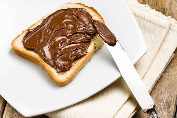 istock Slice of toast with hazelnut chocolate spread on a white plate next to silver knife on rustic wooden table106792090 990480036