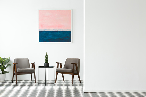 Two grey armchairs, decorative plant on metal table and simple painting hanging on the wall in white open space interior. Empty wall for your product placement