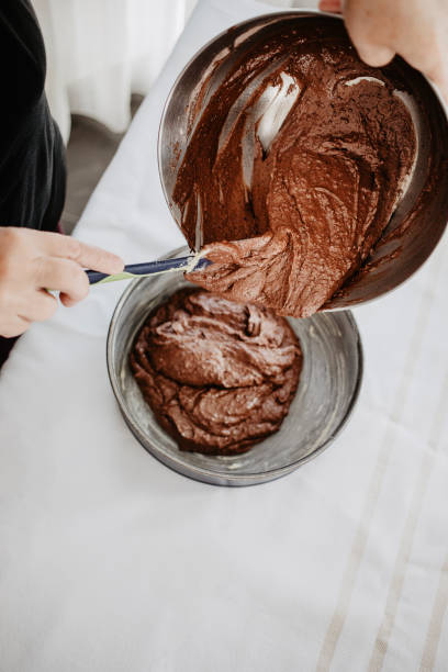 istock Chocolate cake dough being transferred into round-shaped baking tin with silicone spatula. Woman preparing chocolate cake to be baked. White background. 990541908
