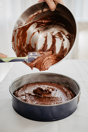 Chocolate cake dough being transferred into round-shaped baking tin with silicone spatula. Woman preparing chocolate cake to be baked. White background.