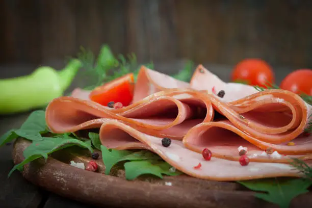 Sliced mortadella on wooden chopping board with vegetables on the background