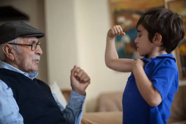 5 years old little boy wearing blue t-shirt showing his biceps to his great grandfather. They are at home. Grandfather is wearing horn rimmed glasses and a hat. He has white hair and mustaches. Shot indoor with a full frame DSLR camera.