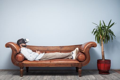 Adult man with VR headset wearing a blue shirt and beige pants lying down on psychiatrist couch. The background is blue wall. His eyes are closed. Shot indoor with a full frame DSLR camera.