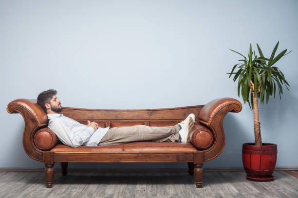 Adult Man Lying Down On Psychiatrist Couch Adult man wearing a blue shirt and beige pants lying down on psychiatrist couch. The background is blue wall. His eyes are closed. Shot indoor with a full frame DSLR camera. leather care stock pictures, royalty-free photos & images