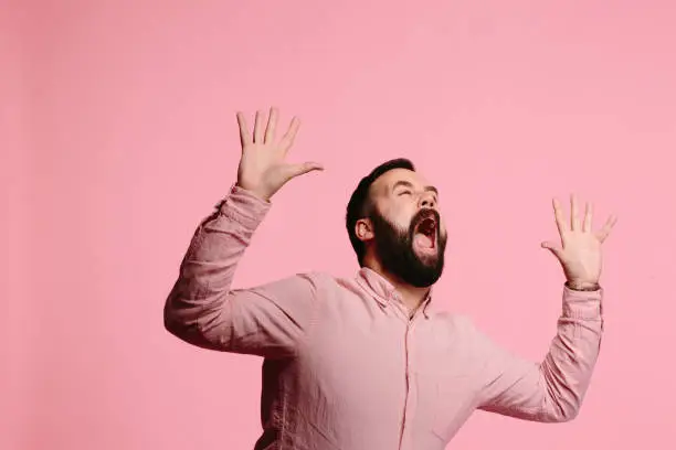 Screaming man with beard and both arms up in the air, mouth wide open, freaking out, isolated on pink background
