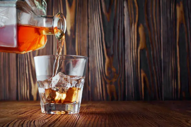 Strong alcoholic drink scotch whisky pour from decanter in old fashion glass with ice cube on wood texture background